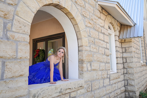 A teen girl, wearing a diamond tiara and diamond heart necklace and royal blue ball gown, leans out from  an arched window of a historic church in Texas. Christmas wreath in the background.