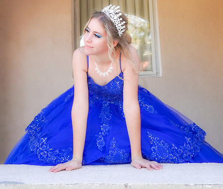 A teen girl, wearing a diamond tiara and diamond heart necklace and royal blue ball gown, looks dreamily off to the side. Close-up of head and shoulders.