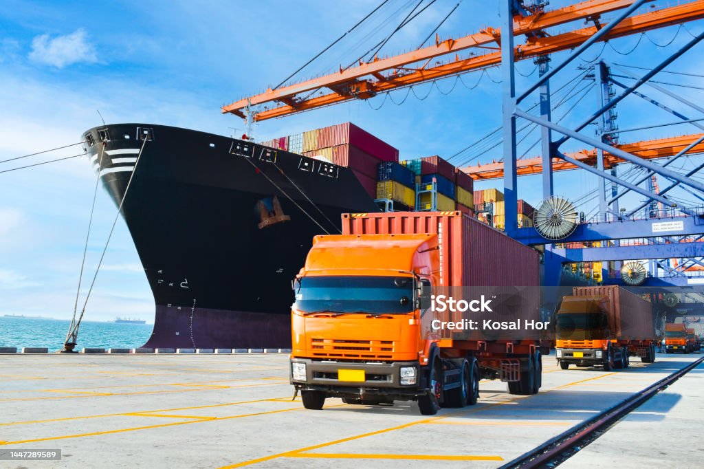 Container shipping, orange color truckload with a container at a harbor. Container port terminal, container delivery. Commercial Dock Stock Photo