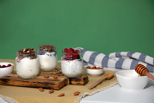 Yoghurts in glass jars with granola, chocolate, honey and nuts on a green background with kitchen towel. The concept of healthy eating. Front view