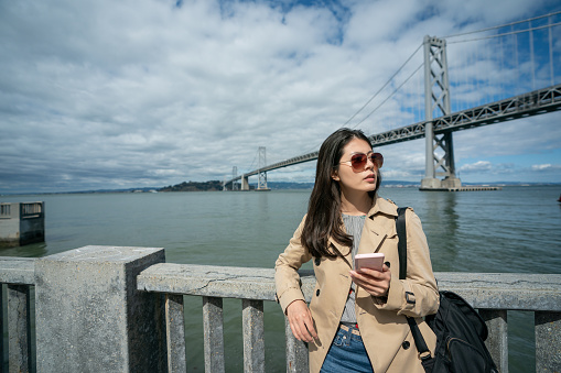 asian chinese woman visitor wearing sunglasses looking into distance with phone in hand while waiting for friend by the railing with Oakland bay bridge at background in san Francisco