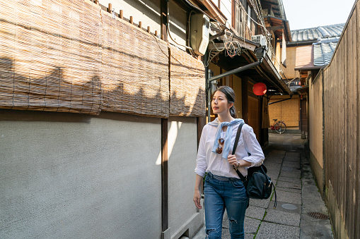 curious asian Japanese woman backpacker looking at traditional bamboo noren curtains on the building wall while walking in an alleyway on Gion Hanamikoji Street in Kyoto japan