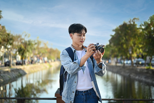 Asian man traveller with backpack taking photo with digital camera while standing on a bridge over a canal.