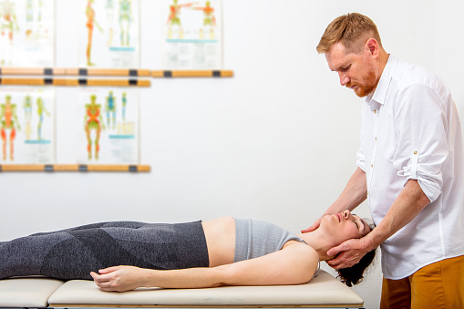 Male osteopath (chiropractor) healing a young woman lying down on a massage bed by touching her head and neck. Can illustrate the concept of Osteopathy, Alternative medicine, Physiotherapy, pain relief. Space for copy.