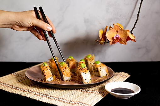 A person holding a sushi roll with the help of black chopsticks, orchid decoration