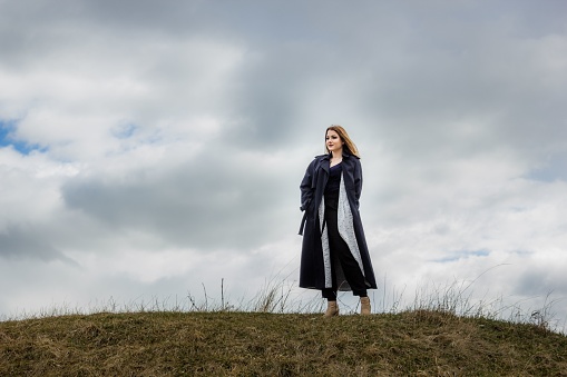 A stylish caucasian woman standing on a grassy hilltop against a cloudy sky