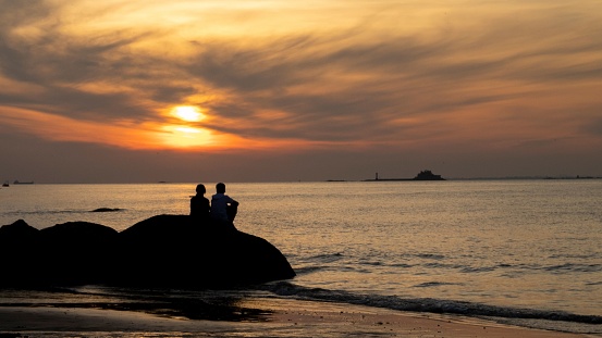A scenery of a silhouette of a couple sitting on a big rock at the beach and admiring the sunset