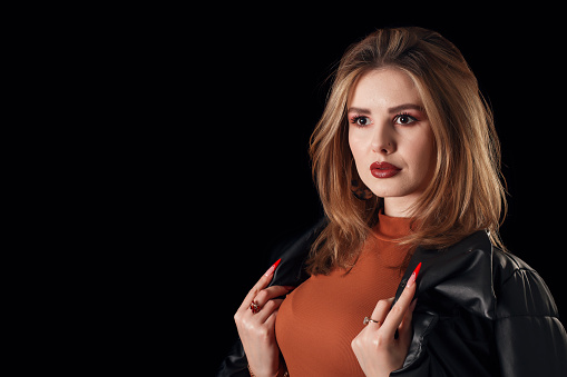 Stunning blond woman in a leather coat poses in front of black background