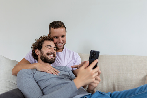 Cute young gay couple video calling their friends in their living room at home. Two male lovers smiling cheerfully while greeting their friends on a smartphone. Young gay couple sitting together.