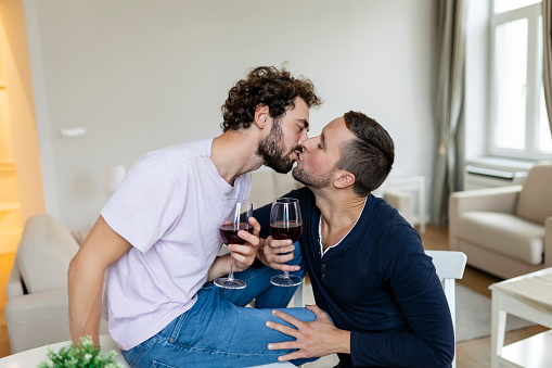 LGBTQ+ couple embracing each other and drinking wine indoors. Two romantic young male lovers looking at each other while sitting together in their living room. Young gay couple being romantic at home.