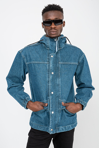 Black male model wears a denim jacket and is in front of a white background. Charismatic black man with his hands in his coat pockets.