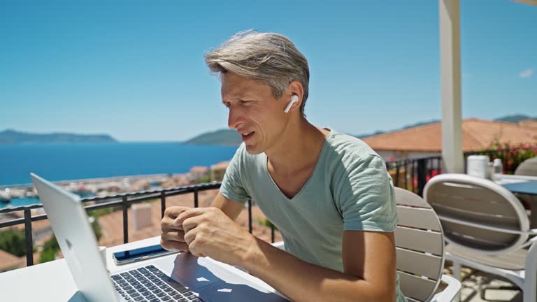 Concentrated skilled freelancer or businessman wearing earphones holding video call, spiking with client or team during summer vacation at sea resort in Turkey. Remote work.