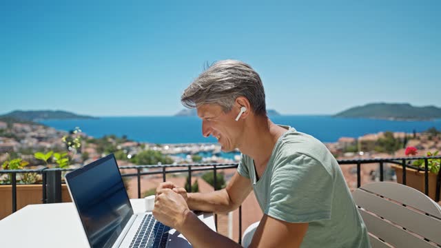 Dolly Zoom Out Imágenes Hombre en auriculares trabajando con computadora portátil al aire libre por hermosa vista azul al mar, hablando por videollamada en línea. Concepto de trabajo y viajes remotos photo