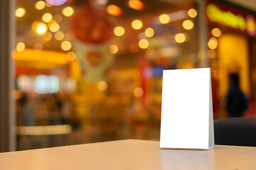 Mock up Label the blank menu frame in Bar restaurant ,Stand for booklets with white sheets paper acrylic tent card on wooden table cafeteria blurred background can inserting the text of the customer.