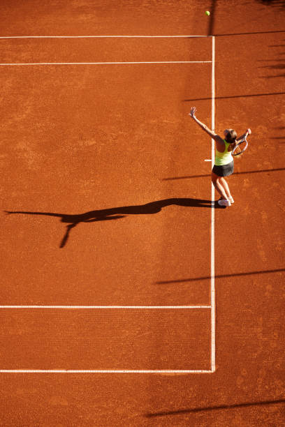 Aerial view of young adult female with shadow serving the tennis ball on clay court stock photo