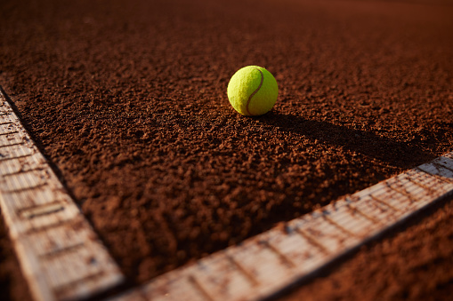 One single tennis ball between two white lines in the corner of a clay court with no people. Copy space,