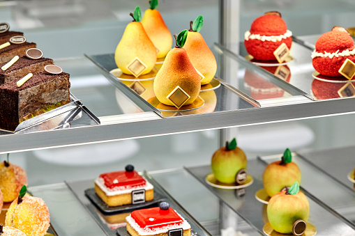 Tempting colorful pastries in shape of pears, apples and peaches, slices of chocolate cake and small cheesecakes topped with fruit jelly and berries on display. Concept of authors confectionery