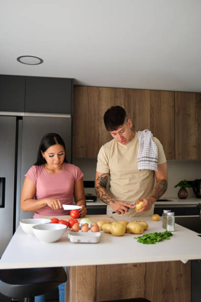 Two young latin people peeling potatoes and cutting tomatoes at kitchen.