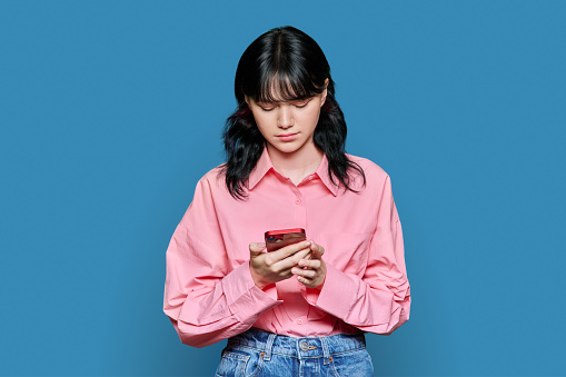 Sad concentrated young female looking at smartphone screen, on blue color background. Teenage student girl reading serious bad news