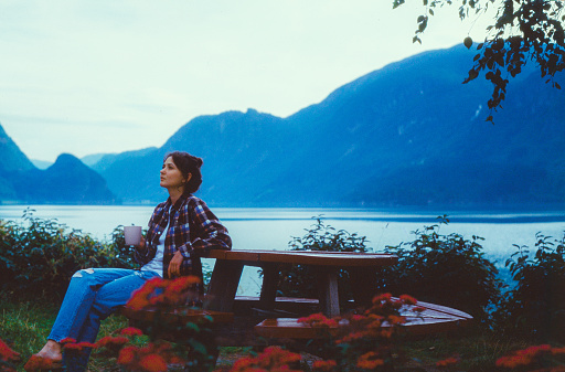 Young Caucasian woman sitting on the bench and drinking coffee on the background of the lake in Norway