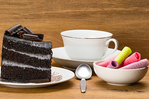 Chocolate sponge cake topping with chocolate tube in white ceramic dish with multicolor coconut wafer tube in white bowl and a cup of coffee on wooden table.