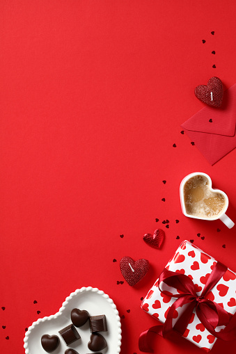 Happy Valentines day concept. Top view table with gift box, heart shaped candy, coffee cup, envelope, confetti on red background. Valentines Day vertical banner template.