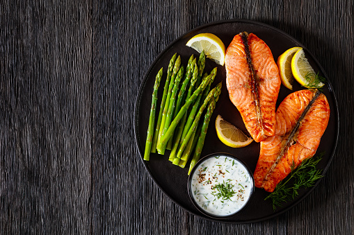 roast salmon steaks with boiled asparagus, fresh lemon and yogurt dill sauce on black plate on dark wood table, horizontal view from above, flat lay, free space