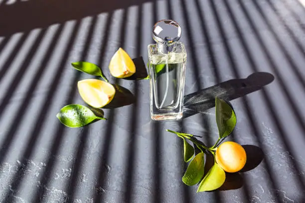 Photo of a clear bottle of cosmetic with a natural citrus aroma stands on a dark table in with parallel shadows from the window blinds.