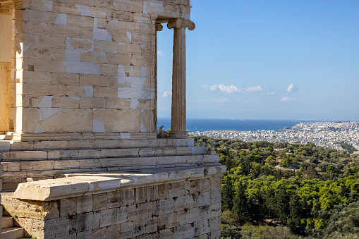 Athens, Greece - October 17, 2022: Temple of Athena Nike at Propylaia, monumental ceremonial gateway to the Acropolis of Athens. In the distance, aerial view of the city and sea with port of Piraeus
