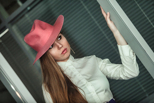 Portrait fashionable young woman model indoors standing at office wall wearing white shirt and red hat, looking away. Stylish beauty lady posing indoor, lifestyle. Fashion style concept. Copy space
