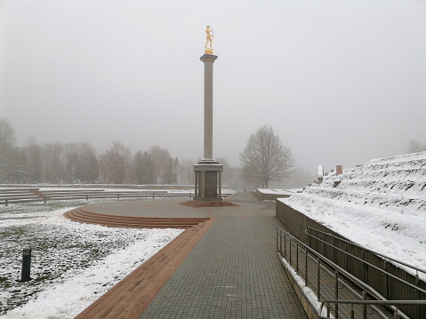 Golden statue in city. Boy statue. Old cemetery in background. Thick fog in distance. Seats near monument. Tiled footpath. Auksinis berniukas Siauliuose.