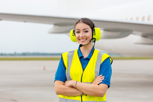 Outdoor shot of young woman standing outside in front of airplane with headphone and smiling at camera. Airport ground crew at work.