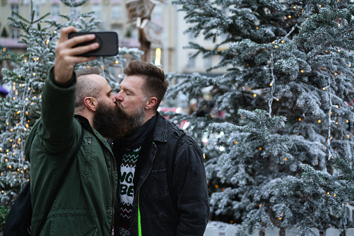 Long beard hipsters homosexuals enjoying Christmas advent, kissing while taking selfie in front of Xmas tree outdoor. Attractive male gay couple celebrating winter holiday season and their love.