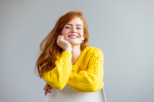 Portrait of young red-haired woman against gray background. Smiling friendly young woman with long red hair wearing spectacles looking at the camera with a vivacious smile against a white interior wall with copy space