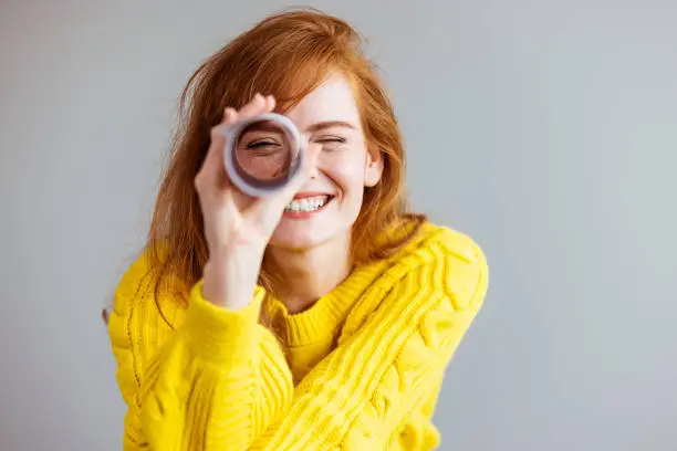 Young woman playing I spy with the camera looking through a rolled cylinder of paper with one eye and a happy smile. Redhead woman looking through paper telescope