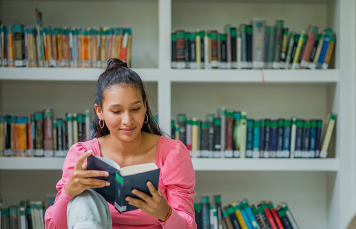 Female college student sitting in library by the bookshelves and reading book