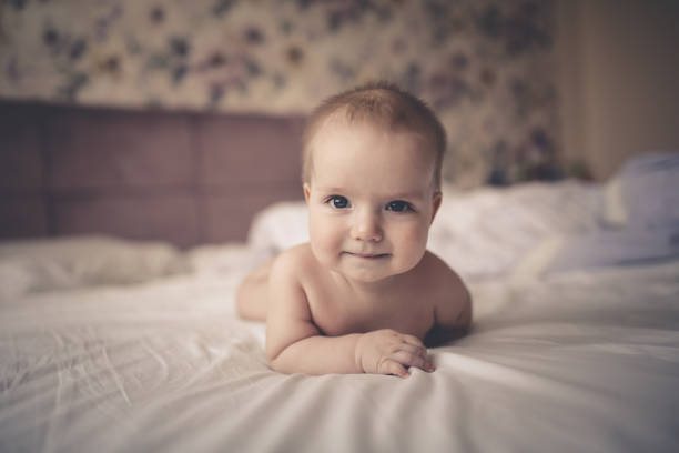 baby lying on his stomach on bed in bright real Adorable baby 6 months old lying on his stomach on a bed in a bright real interior, a childhood end. looks into the frame half dozen babies stock pictures, royalty-free photos & images