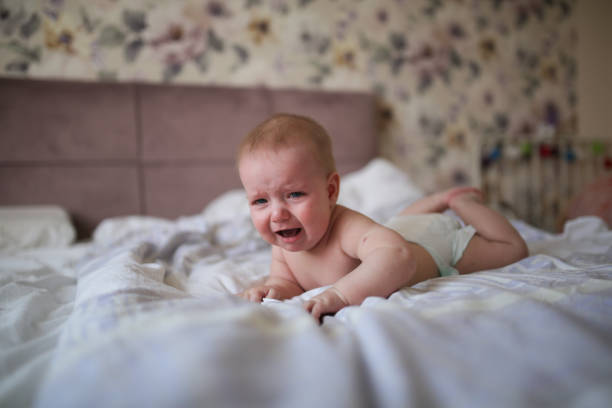 charming baby, 6 months old, crying,on his stomach charming baby, 6 months old, lies crying on his stomach on a bed in a bright, real interior, a childhood end, and teeth are cut, colic of newborns. half dozen babies stock pictures, royalty-free photos & images