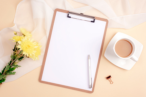 Desktop with blank clipboard, pen, coffee cup and yellow chrysanthemum flowers on light beige background. Top view, flat lay, mockup.