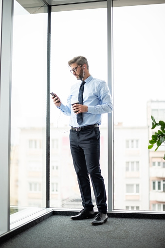 Full length of businessman using smartphone and earphones while standing at the office by the window. Professional man wearing shirt and tie.