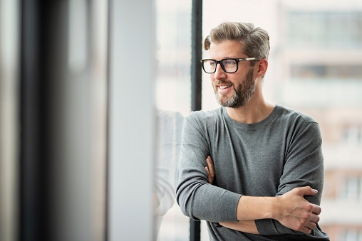 Portrait of confident businessman wearing glasses and sweater while standing at the office and looking out the window.