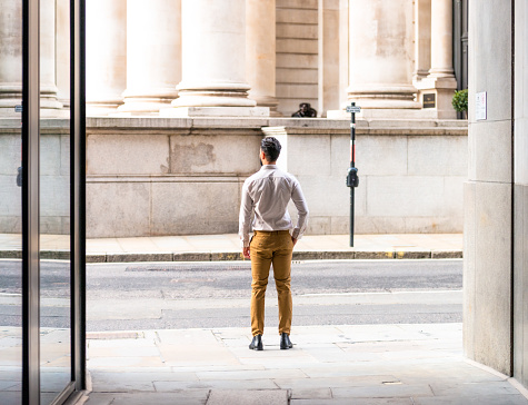 Rear view of a young man standing on the street outside a business in the City of London, the capital's main financial district.

Model released image.