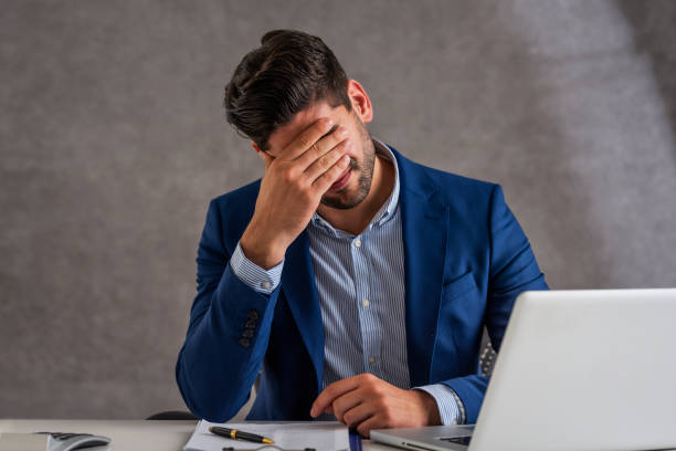 Businessman suffering from strong pain in his head while sitting at office desk Shot of stressed businessman sitting at desk in office with laptop. Professional man suffering from migraine. head in hands stock pictures, royalty-free photos & images