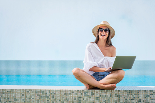 Attractive woman wearing sunglasses and straw hat while relaxing at poolside. Happy female using her laptop.