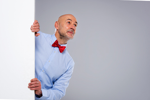 Studio shot of surprised face man looking up while standing behind an empty white board. Isolated grey background. Copy space.