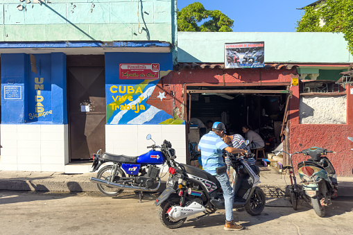 Santa Clara, Cuba - December 6, 2022: People waiting outside an electric bicycle repair shop. A bread-making facility to the left with a propaganda slogan reading \