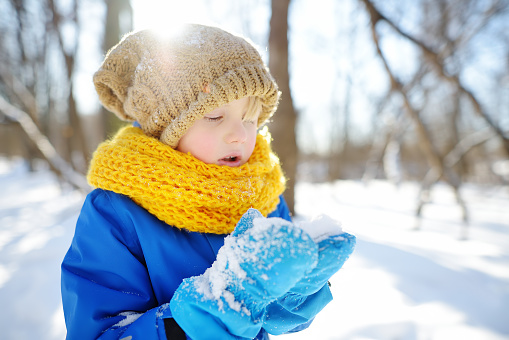 Little boy blowing snow from his hands. Child enjoy walking in the park on snowy day. Baby having fun during snowfall. Outdoor winter activities for family with kids.