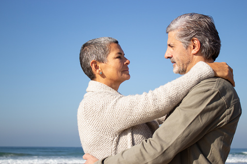 Gray-haired senior couple hugging at seashore on sunny autumn day. Short-haired middle-aged lady and bearded senior man looking at each other. Side view. Relationship, love, traveling concept
