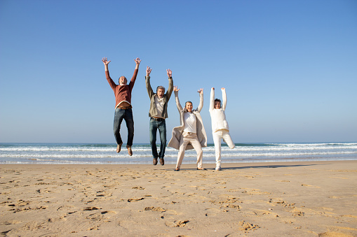 Excited senior friends having fun at beach on sunny autumn day. Four people laughing and jumping with raised arms while having great vacation time at seaside. Vacation, joy, friends concept