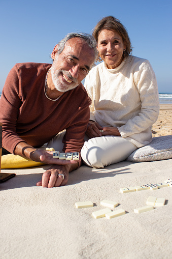 Happy senior couple playing dominoes at seashore on sunny day. Smiling bearded man looking at camera and showing several domino tiles on his palm. Joyful blonde woman sitting behind. Leisure concept
